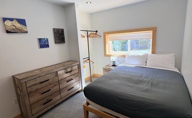 Clean private hostel room with a queen bed, wood dresser, open clothes rail, and small mountain paintings on the wall